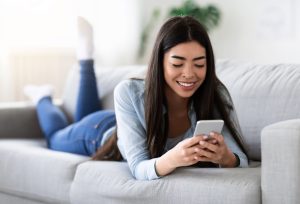 Useful App. Korean Girl Using Smartphone While Relaxing On Sofa At Home